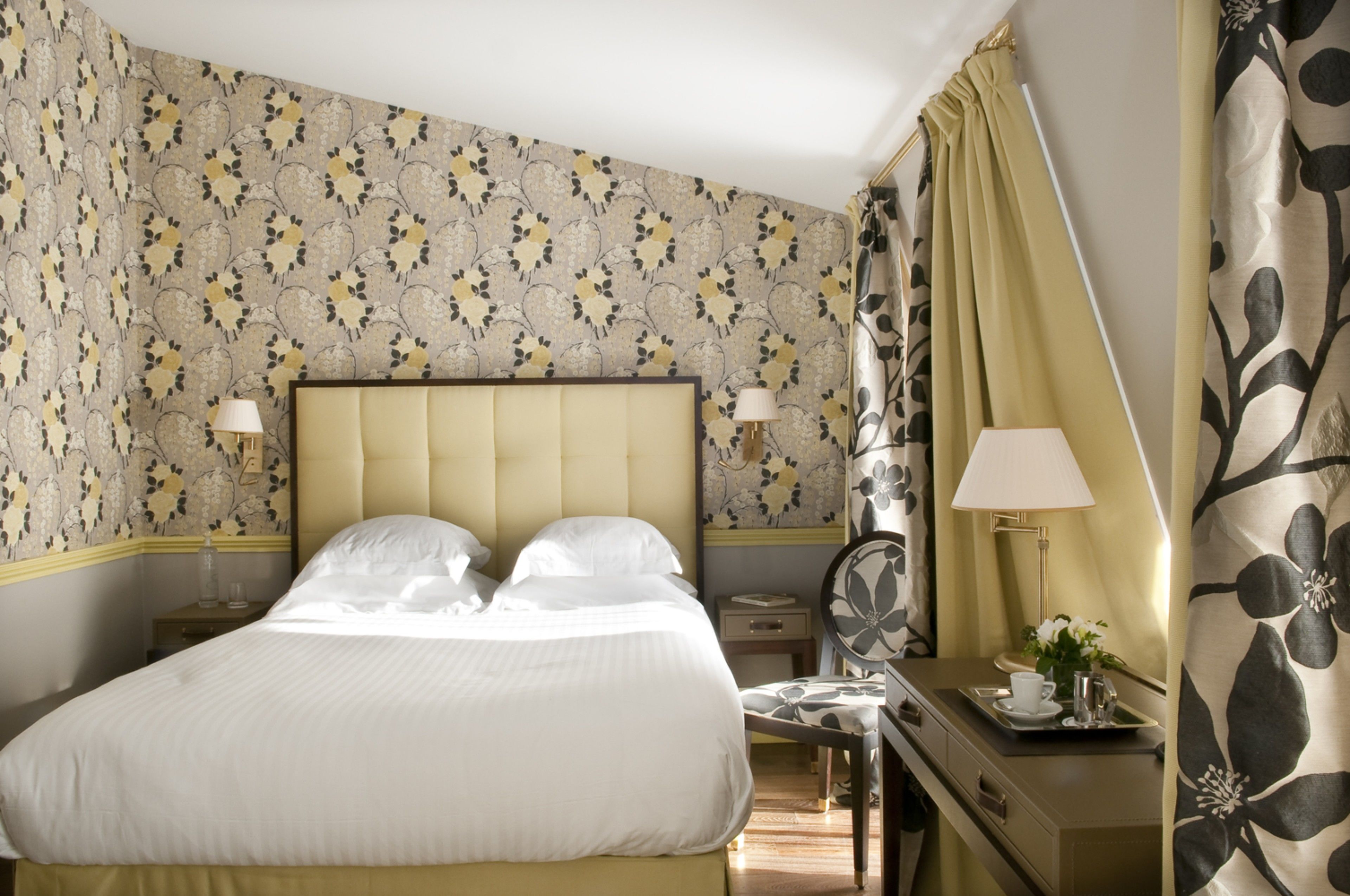 A neatly made bed with a headboard, a bedside table with a lamp, and a tray with a cup and flowers.