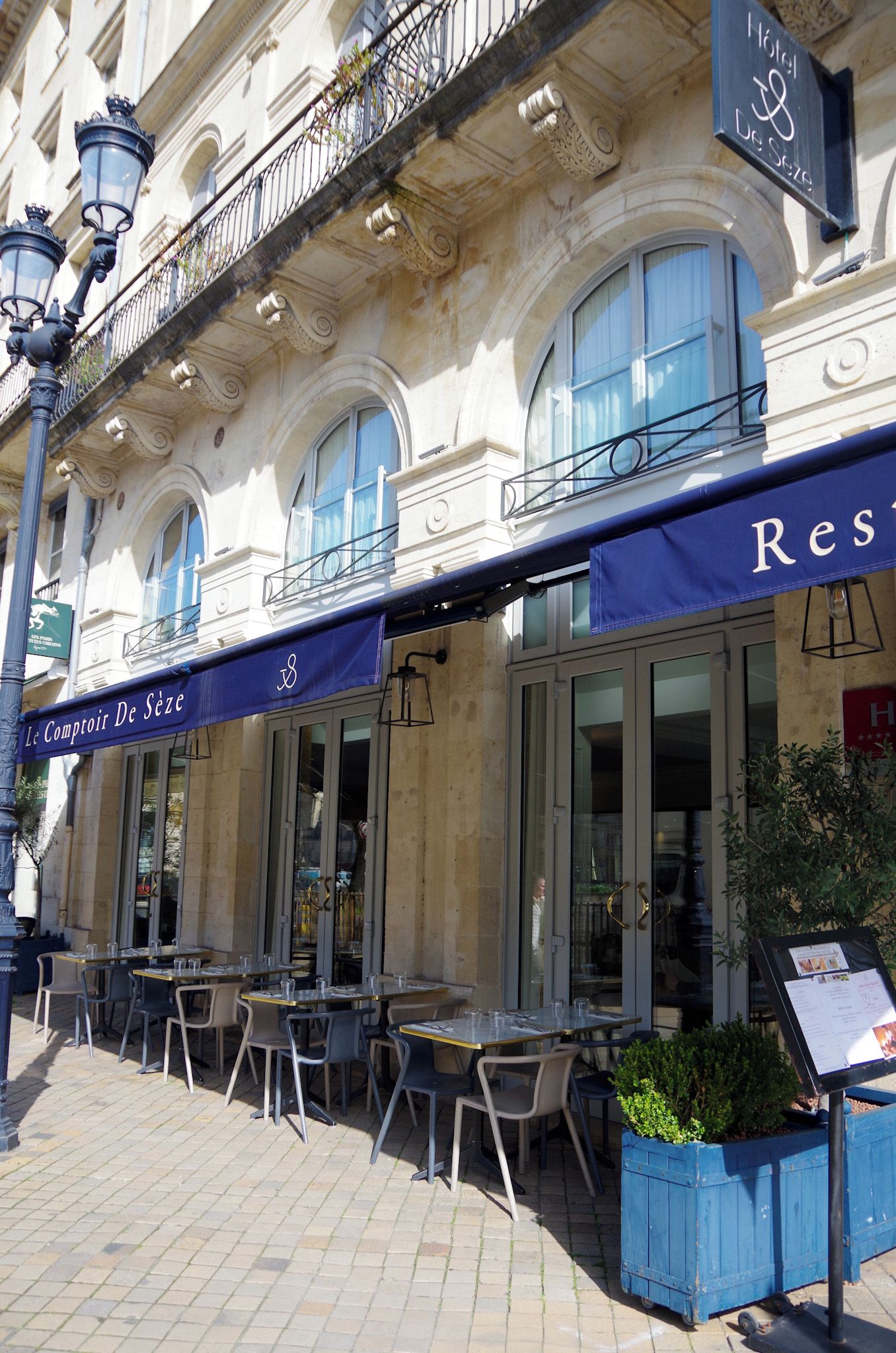 A cafe with outdoor seating, a sign reading 'Le Comptoir De SÃ©ze,' and a building with classical architecture.