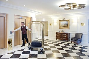 Un vestíbulo de hotel con piso a cuadros, un carrito de equipaje, un tocador de madera y un cuadro enmarcado en la pared.