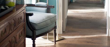 A room with a wooden dresser, a chair, a lamp, and a view into another room with a bed and a chair.
