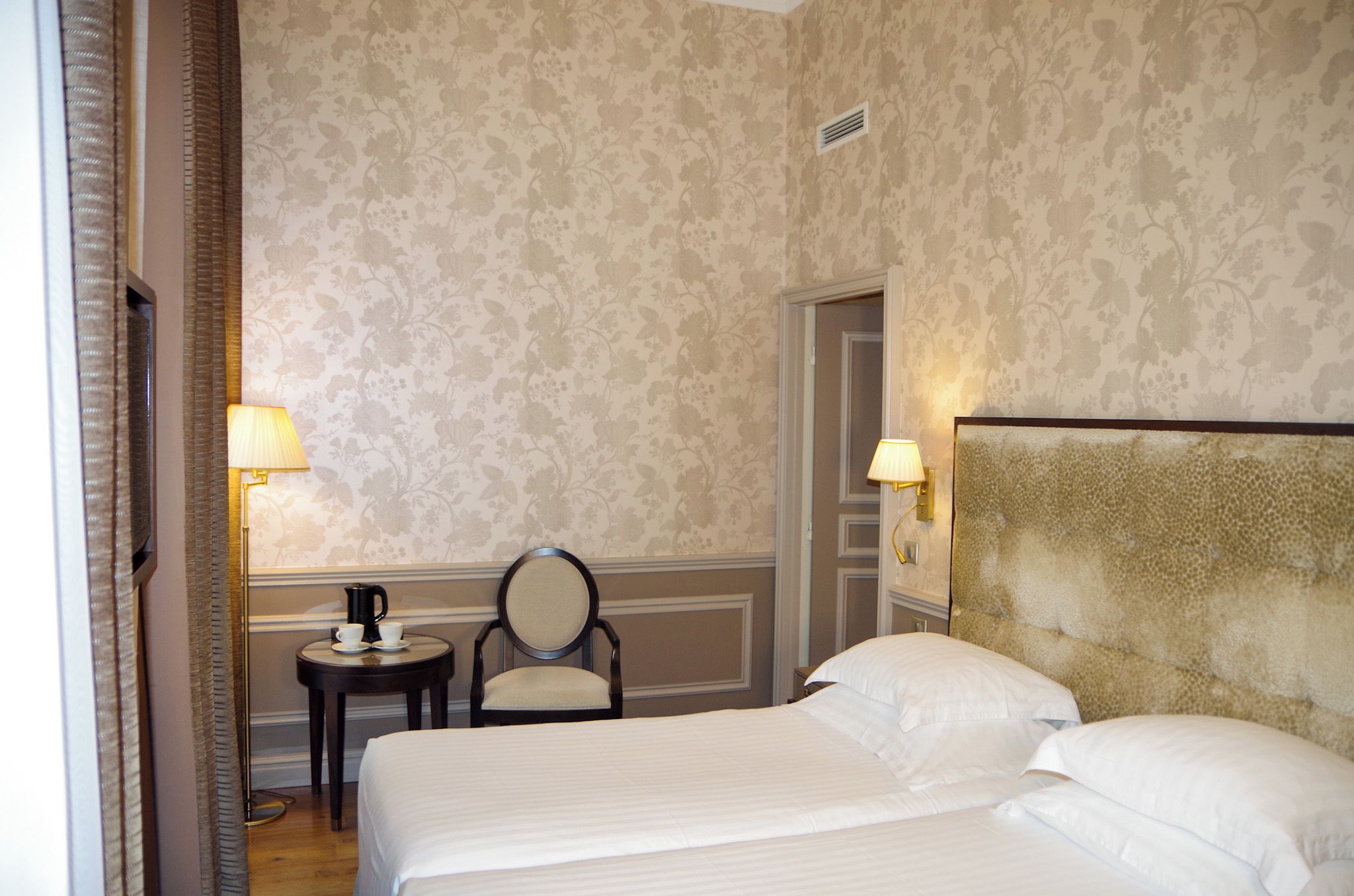 Une chambre d’hôtel avec un grand lit, deux lampes de chevet, une chaise, une petite table avec une cafetière et une tête de lit décorative.