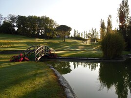 Un campo de golf con un puente sobre un estanque, rodeado de árboles y un césped cuidado.