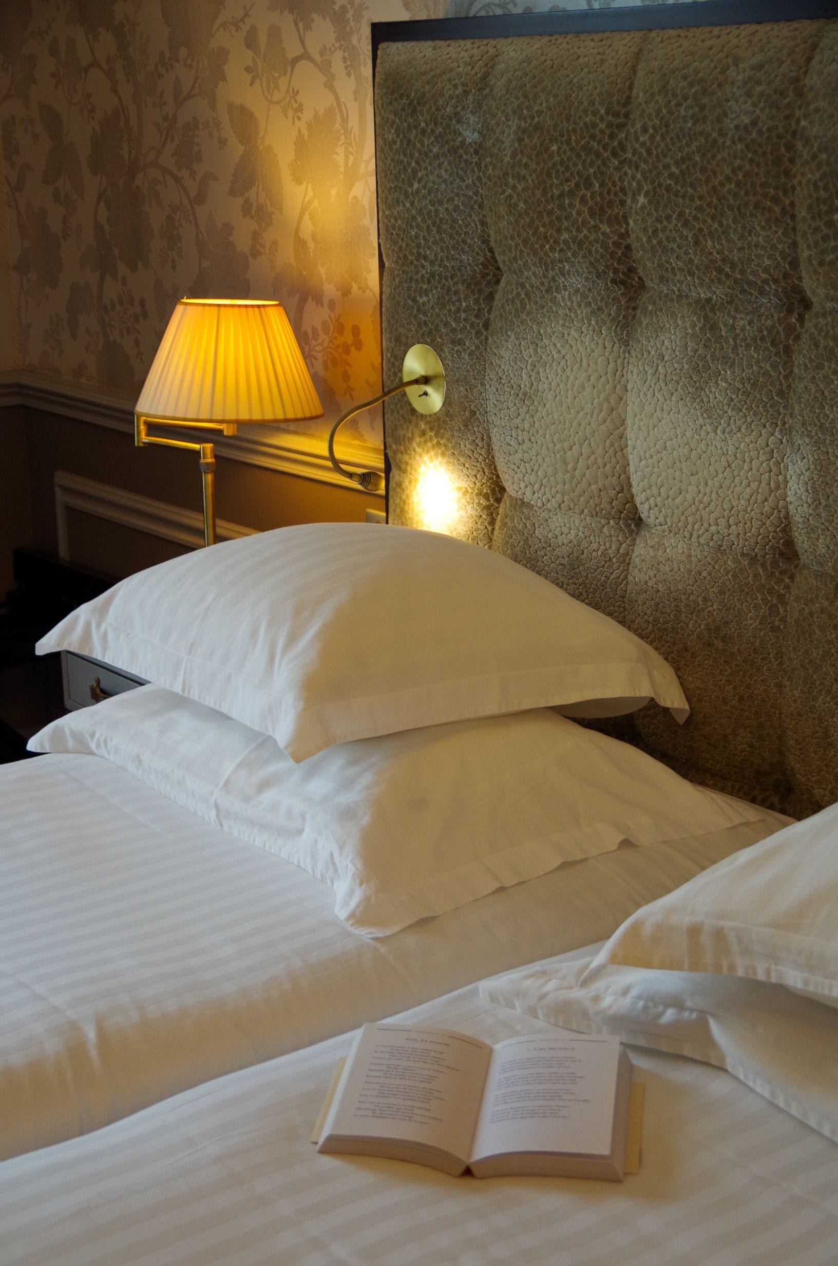 Una cama bien hecha con sábanas y almohadas blancas, una lámpara de noche y un libro sobre la cama.