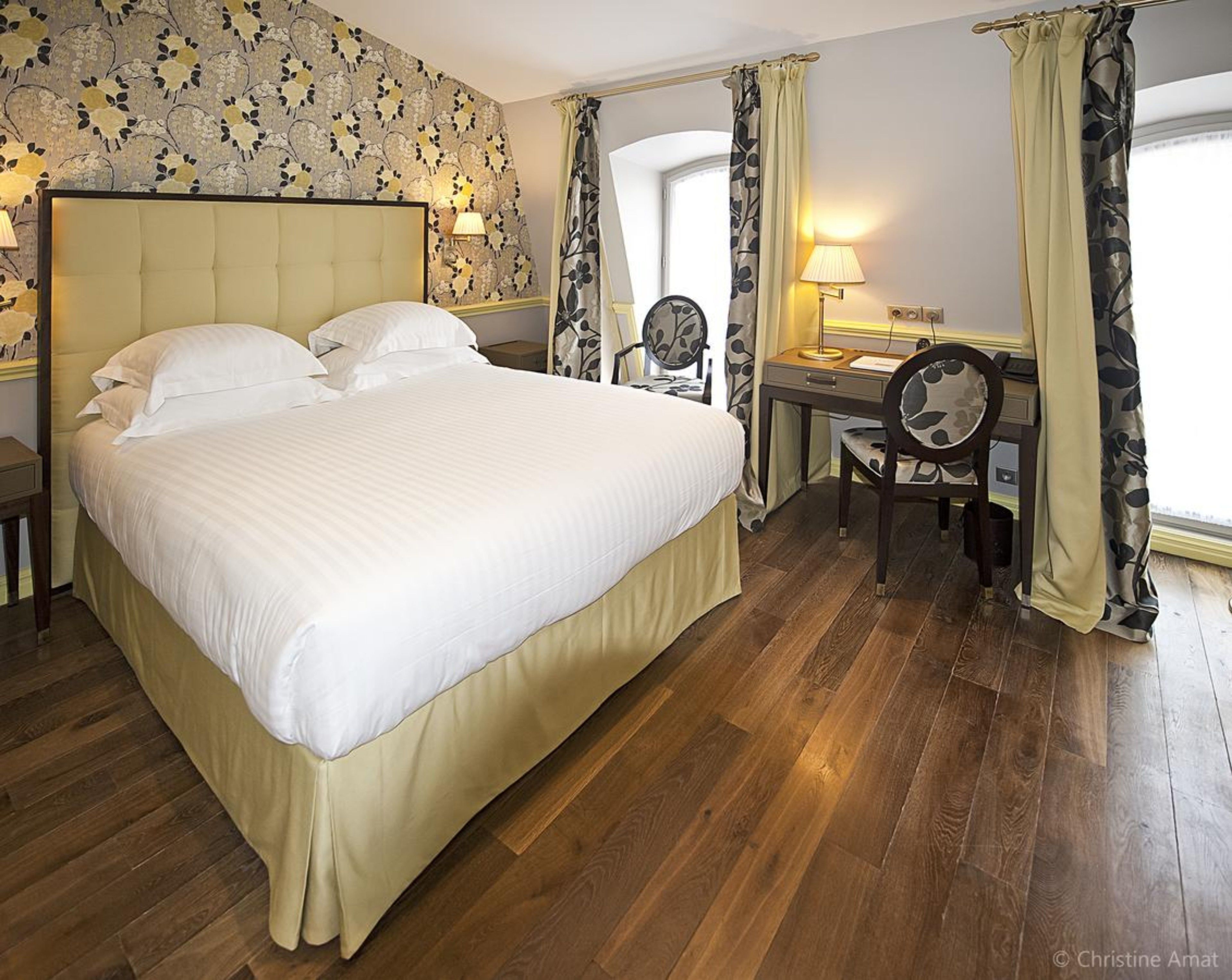 Une chambre d’hôtel avec un grand lit, un bureau avec une chaise, une lampe et une fenêtre avec des rideaux.
