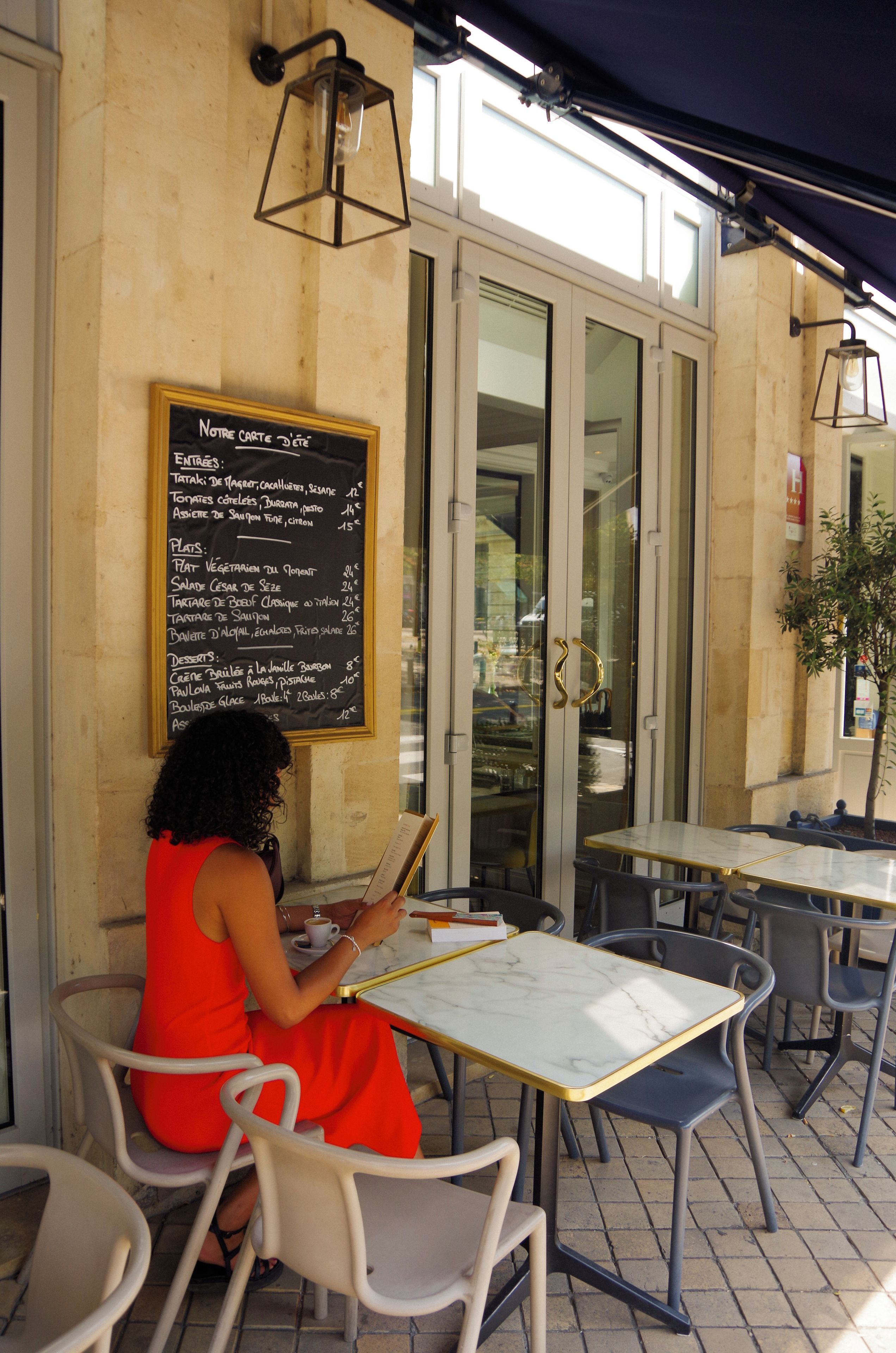 A woman seated at an outdoor cafe with a menu board on the wall.