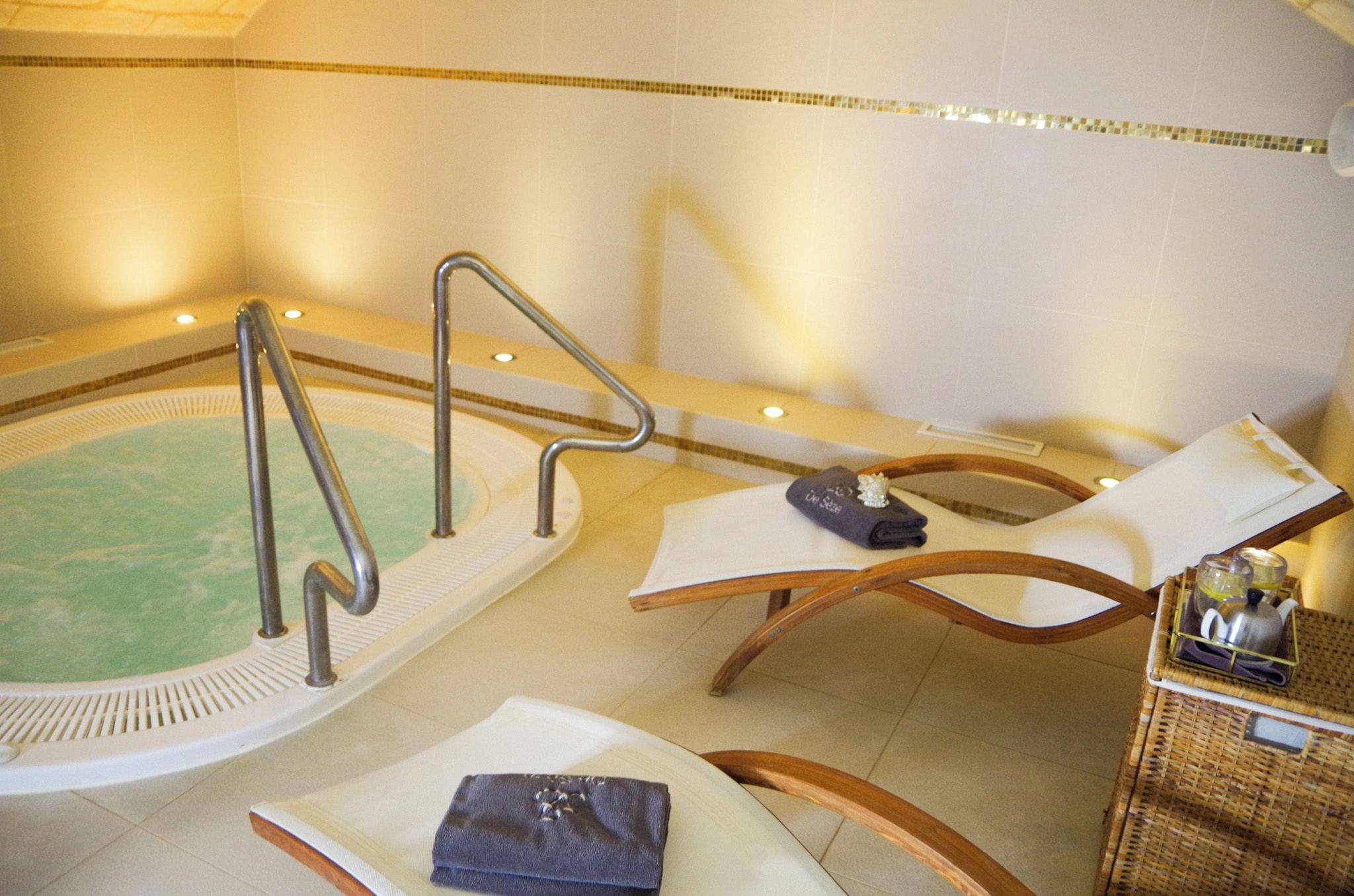 Zona de spa con una bañera de hidromasaje, asientos y una pequeña mesa con una toalla y una botella.