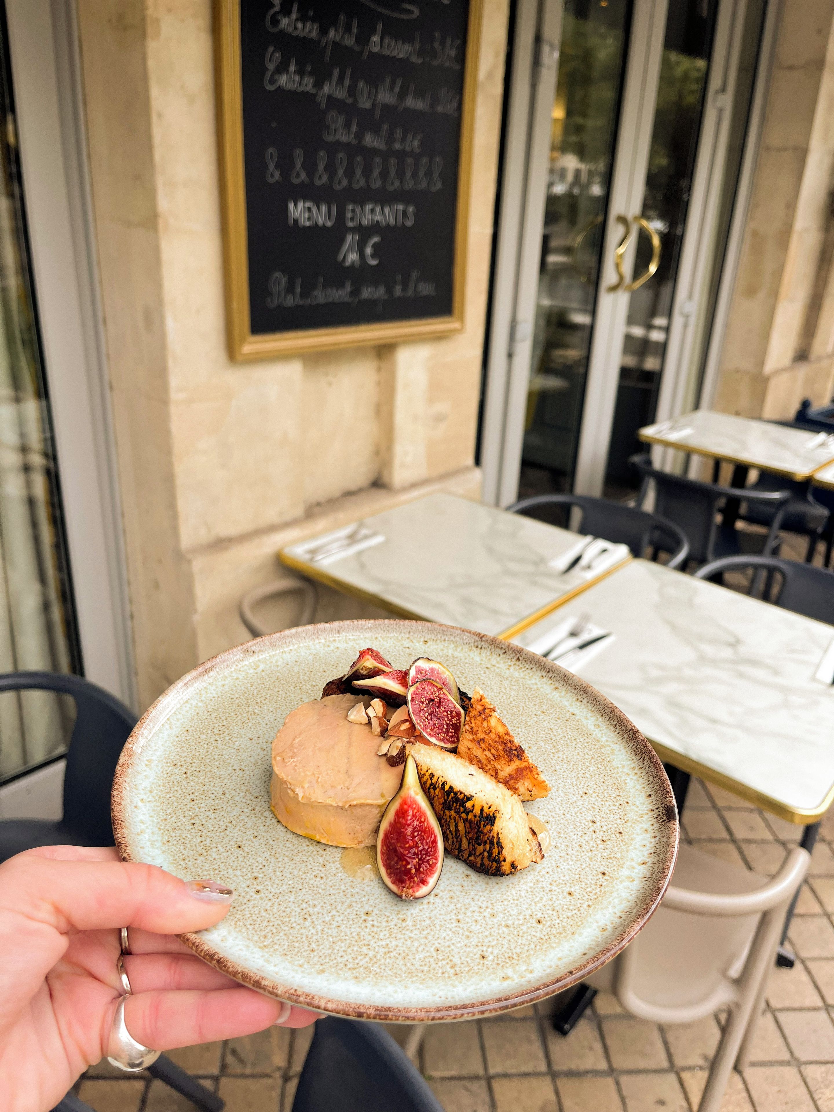 A person holding a plate with a dessert, a menu board, and outdoor seating.