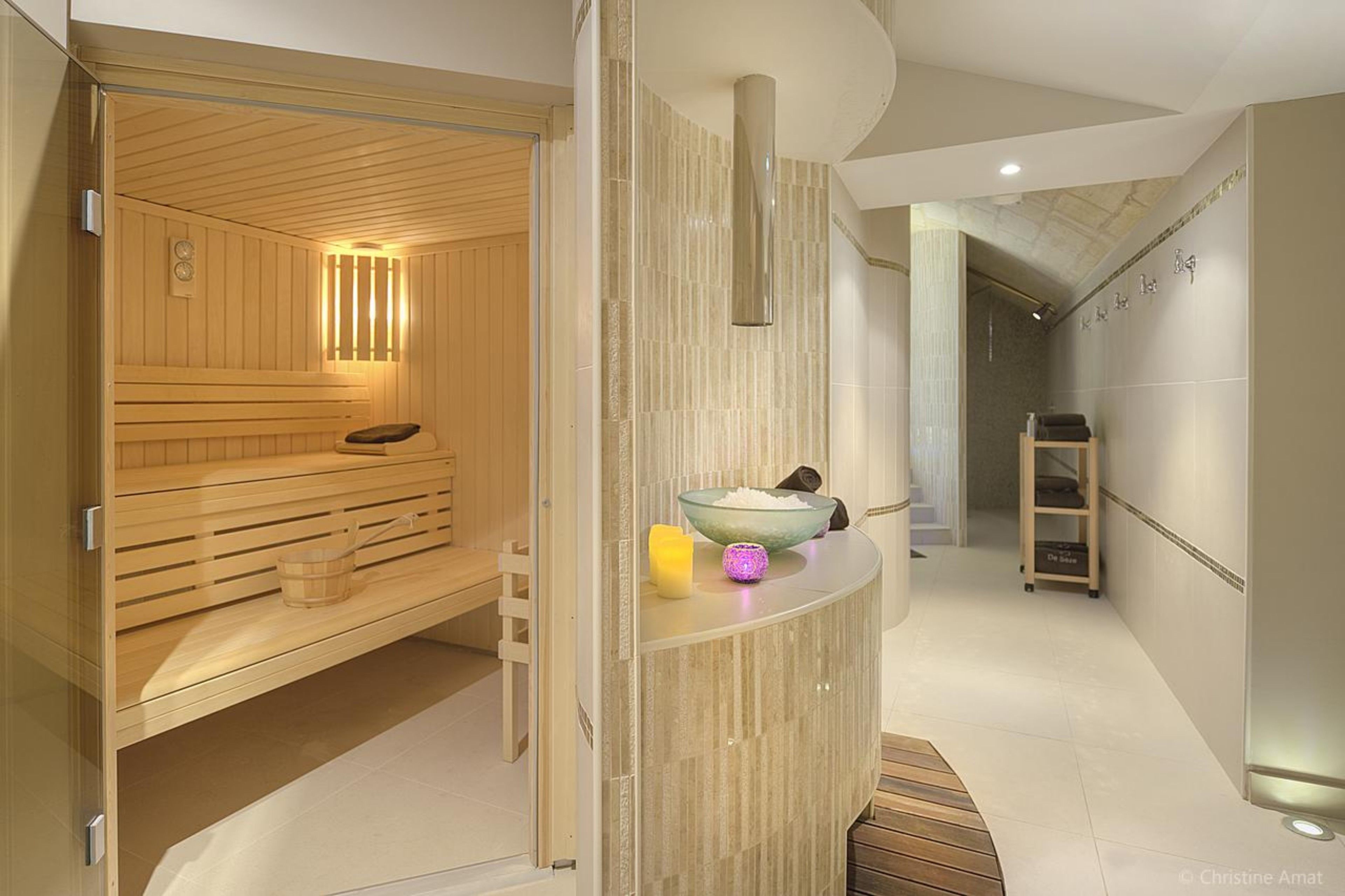 Moderni spa-huone, jossa on puinen sauna, lasiseinällä erotettu hieronta-alue sekä vastaanottotiski, jossa on kulho ja kynttilä.