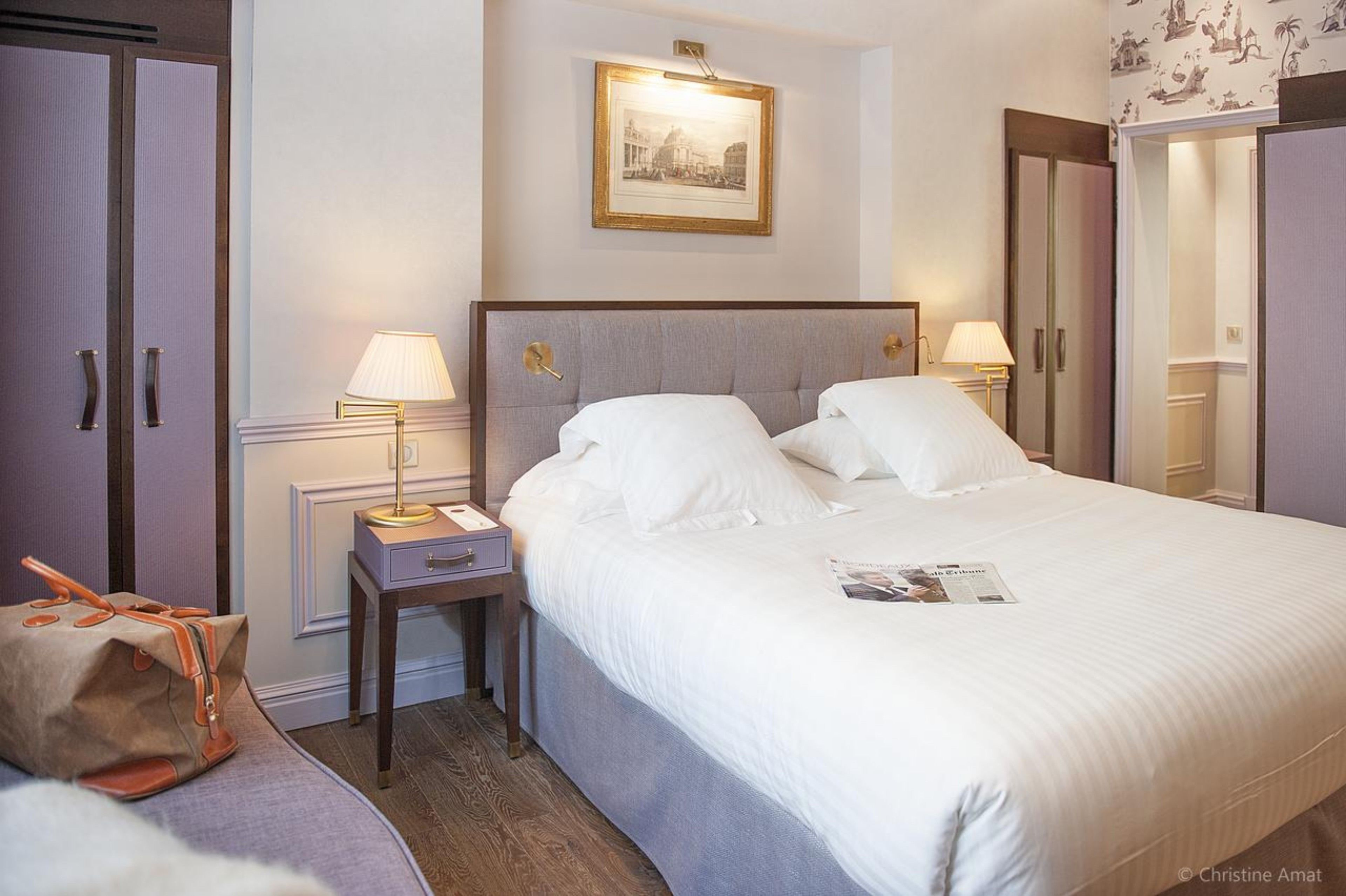 A hotel room with a large bed, two bedside lamps, a nightstand, a framed picture on the wall, and a suitcase on the floor.
