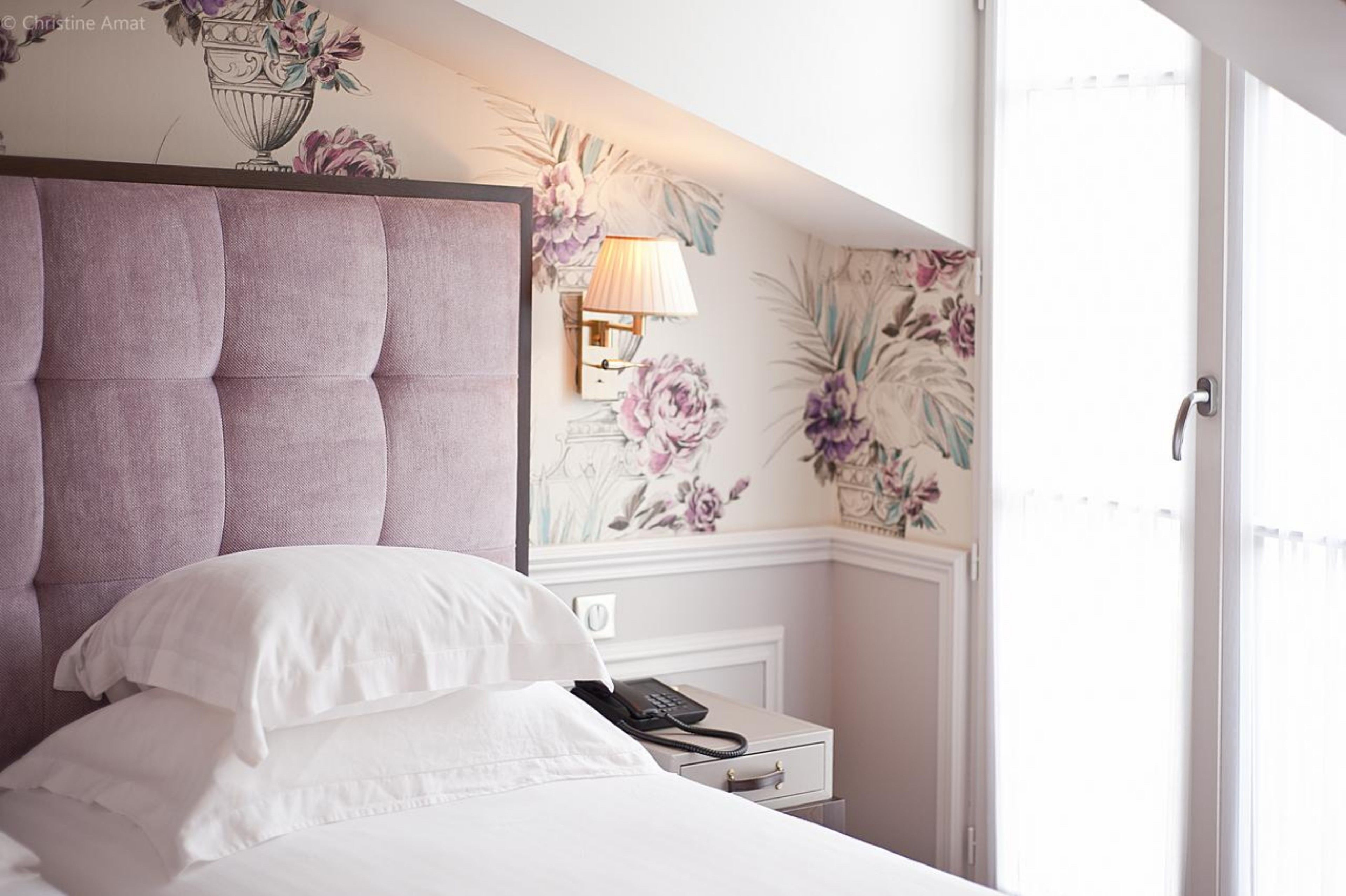 A bedroom with a tufted headboard, a bedside lamp, a floral wallpaper design, and a white nightstand with a phone.