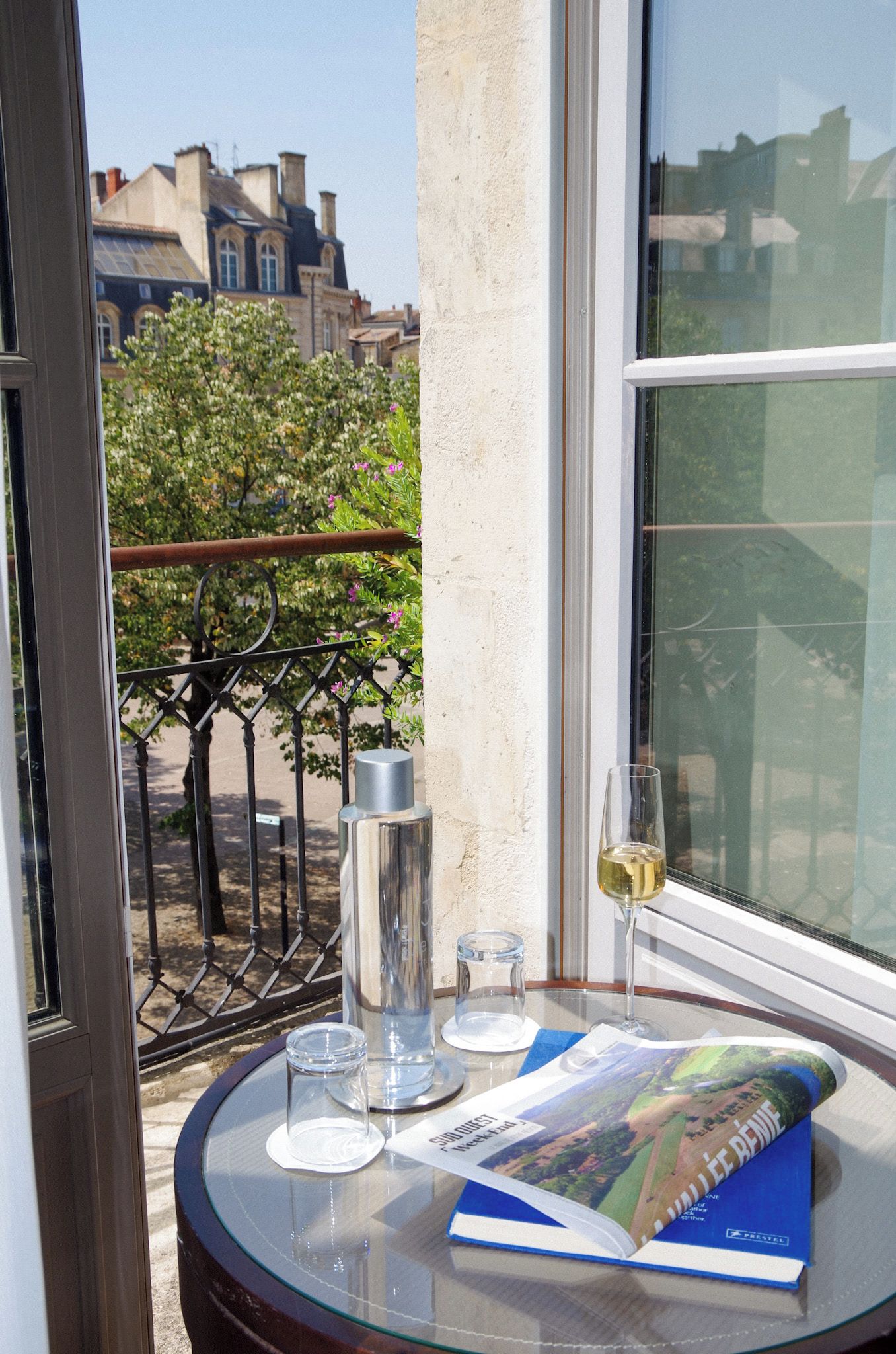 A glass table with a wine glass, a bottle, and a book on a balcony with a view of buildings and trees.