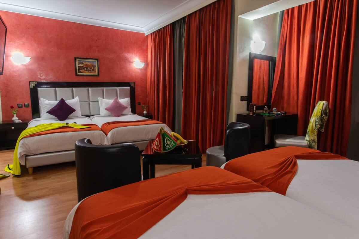 A hotel room with two beds, red curtains, a headboard, a bedside table with a lamp, and a small table with a vase.