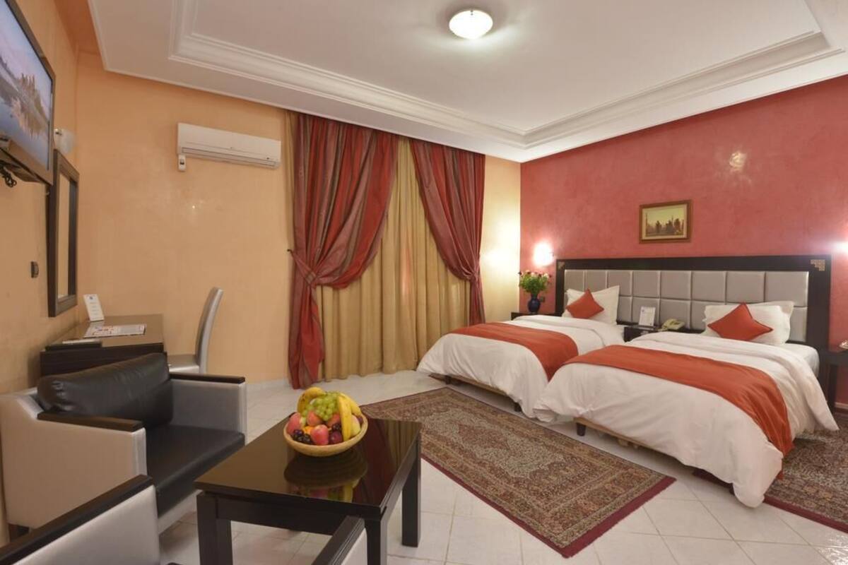 A hotel room with two beds, a desk with a fruit bowl, a chair, and a large window with curtains.