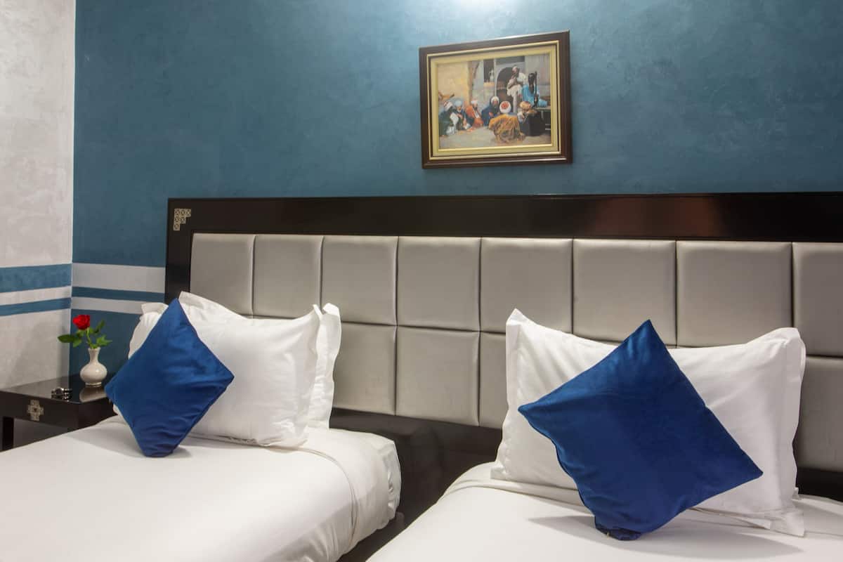 A hotel room with two beds, each with white bedding and blue pillows, a dark headboard, and a framed painting on the wall.