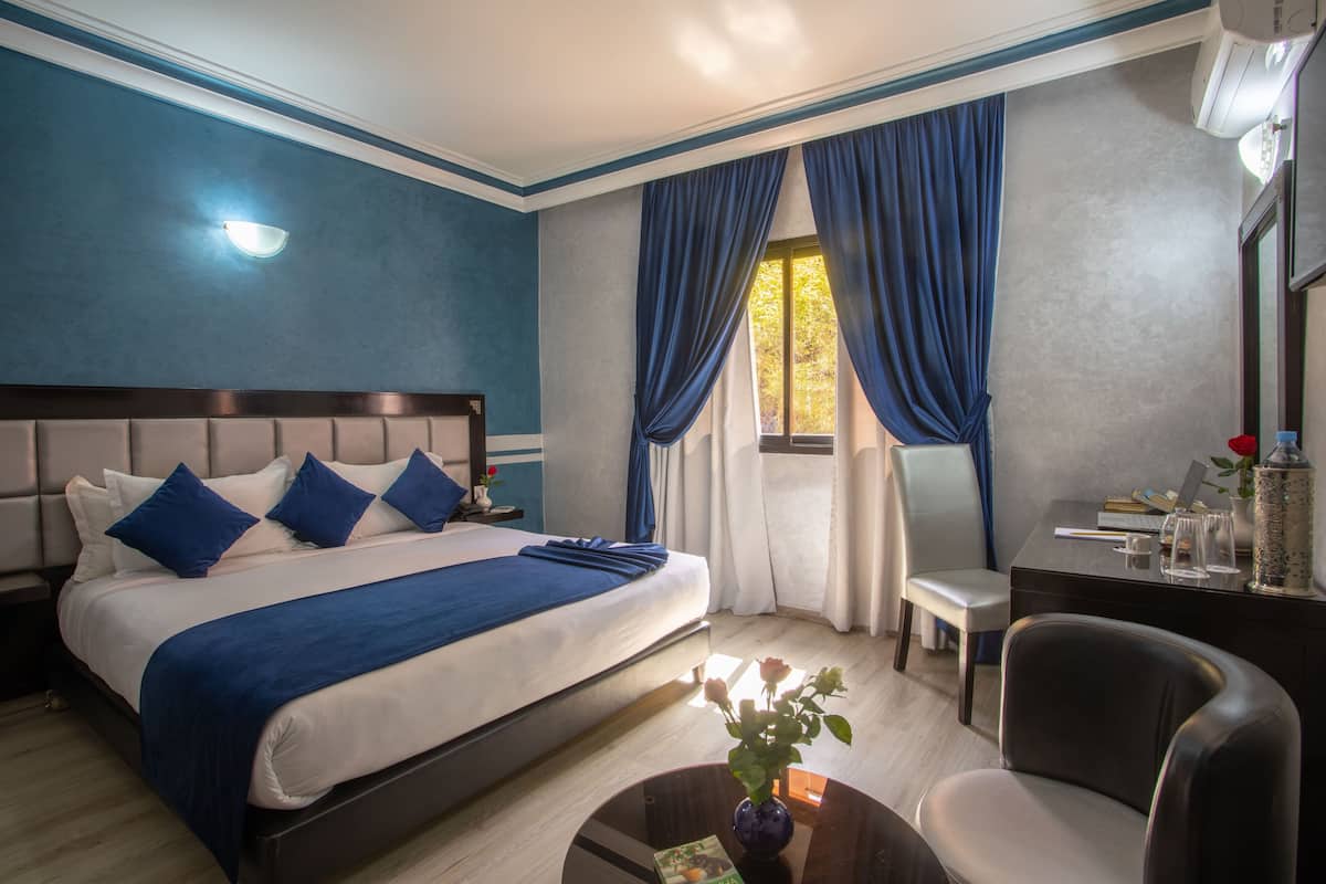 A hotel room with a large bed, a desk, a chair, and a window with blue curtains.