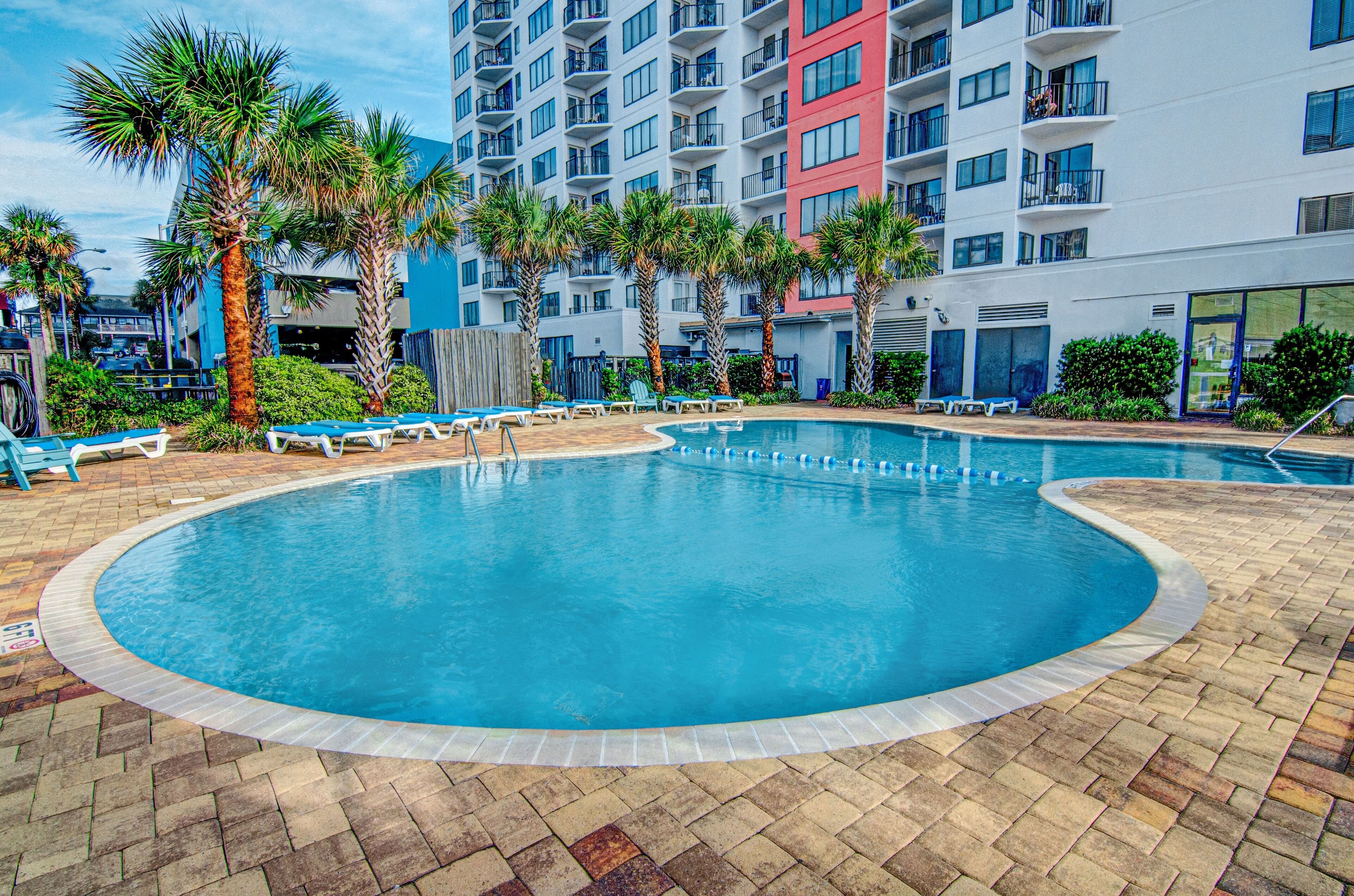 A swimming pool with lounge chairs and palm trees, surrounded by a multi-story building with balconies.