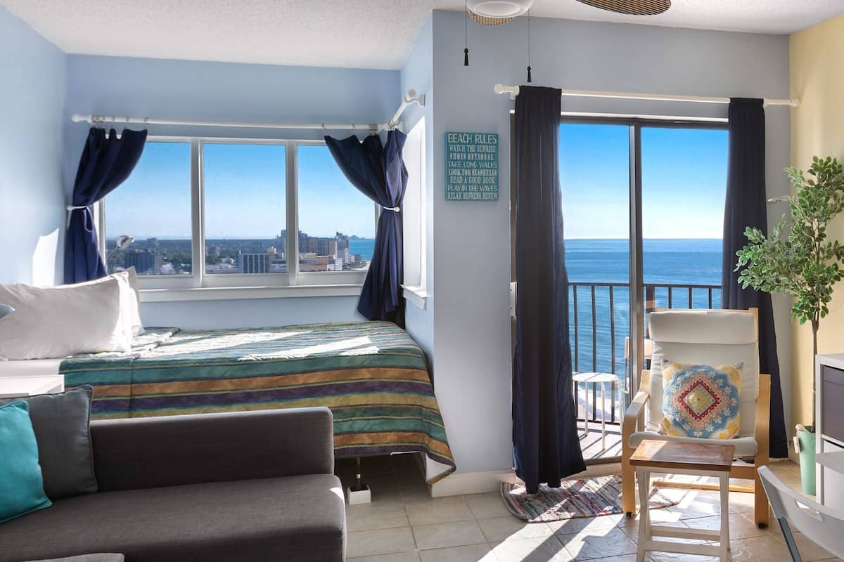 Una habitación con cama, sofá, una mesita, una silla, ventana con cortinas y vista al mar.