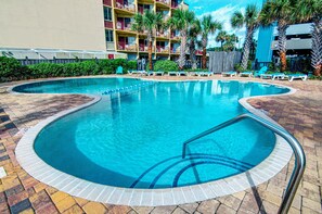 A swimming pool with a tiled deck, lounge chairs, palm trees, and a multi-story building in the background. - Palace Resort (Myrtle Beach)