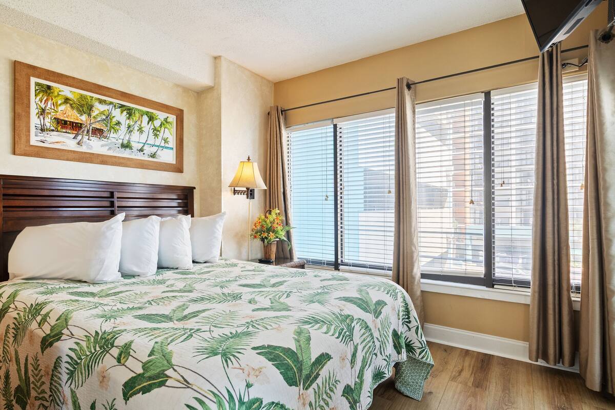 Una habitación de hotel con cama, mesita de noche con lámpara, cuadro de una playa y ventanas grandes con persianas.