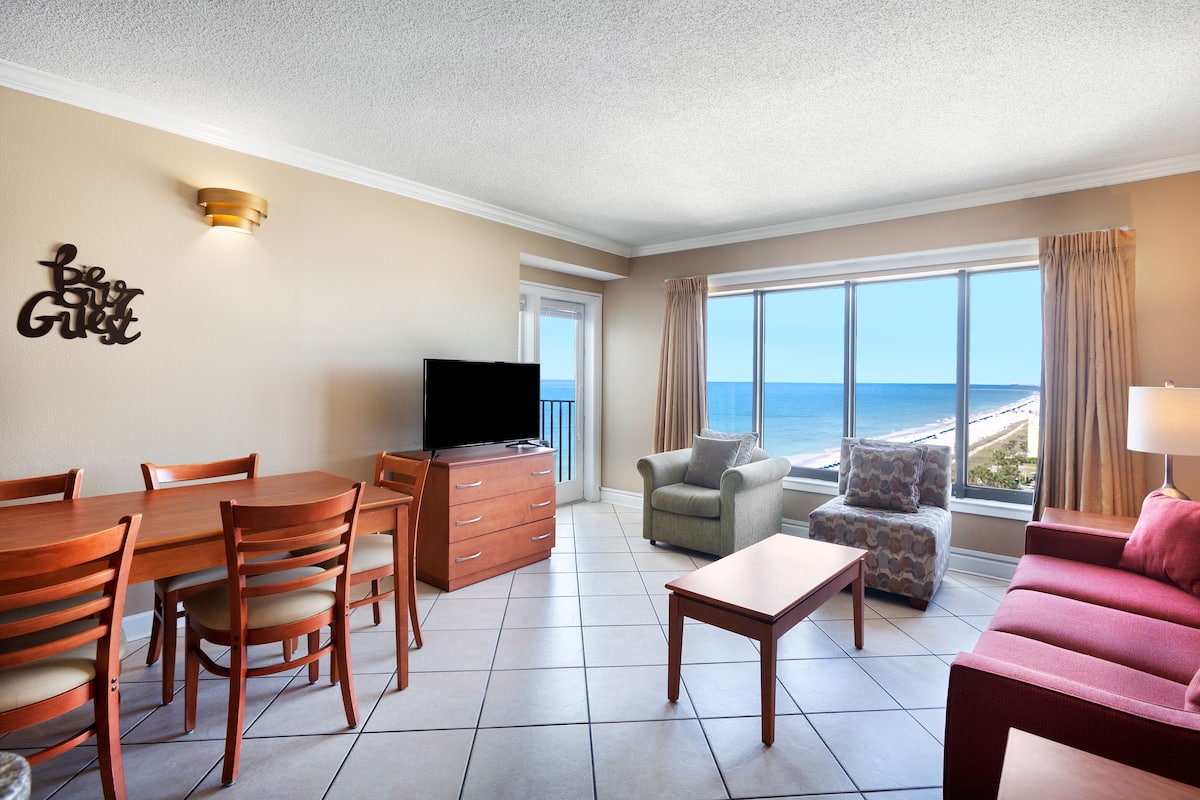 Habitación de hotel con zona de comedor, televisión, sofá y vista a la playa.