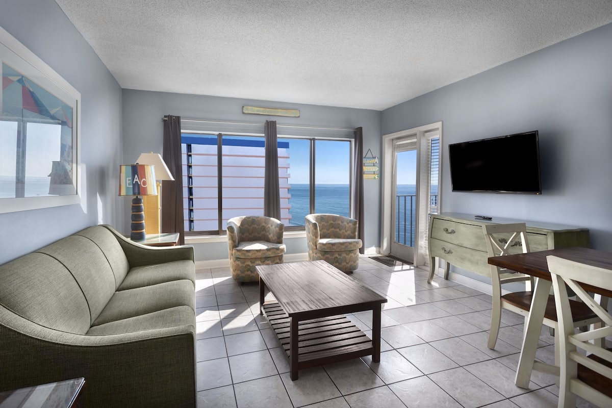 Una sala de estar con sofá, dos sillones, una mesa de centro, televisor y un balcón con vista al mar.