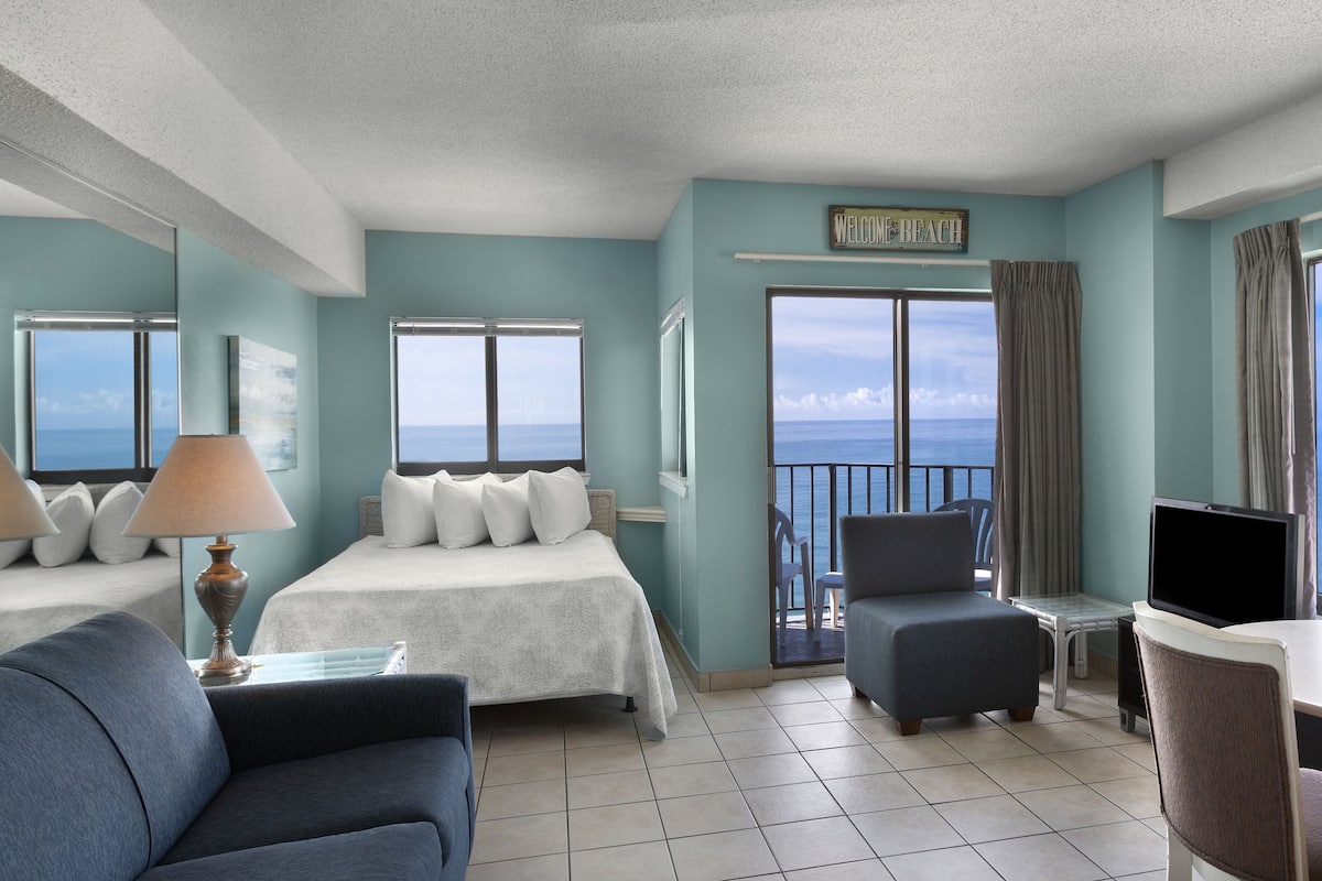 Una habitación de hotel con cama, sofá, televisión, balcón con vistas al mar y un cartel que dice 'Welcome Beach'.