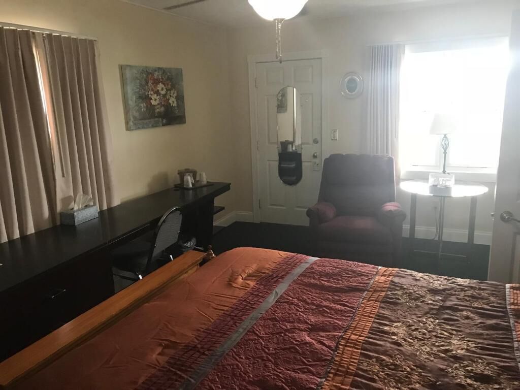 Et hotelværelse med seng, skrivebord, stol, lille bord, lampe, et vindue med gardiner og en dør med spejl.
