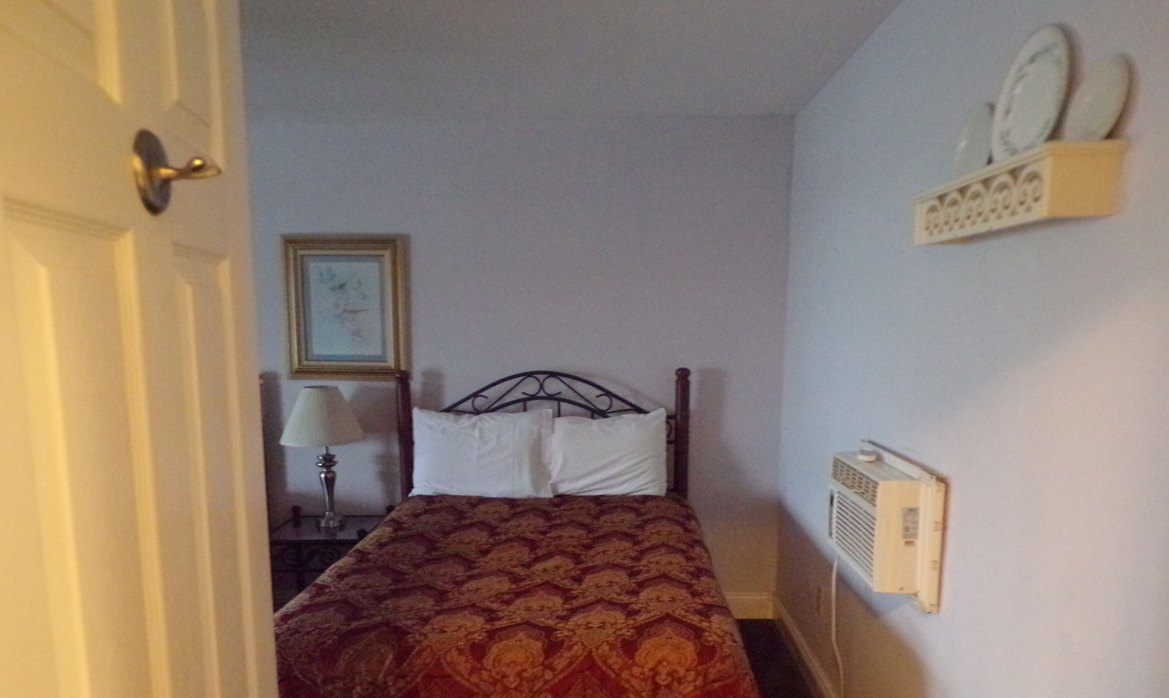 A bedroom with a bed, a bedside table with a lamp, a wall-mounted heater, and a shelf with plates.