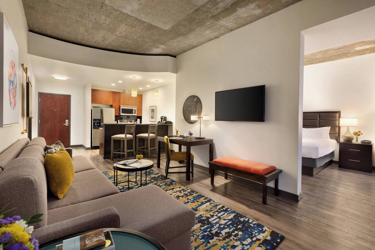 A modern hotel room with a sofa, a small round table, a bench, a desk, a flat-screen TV, a bed, and a kitchen area in the background.