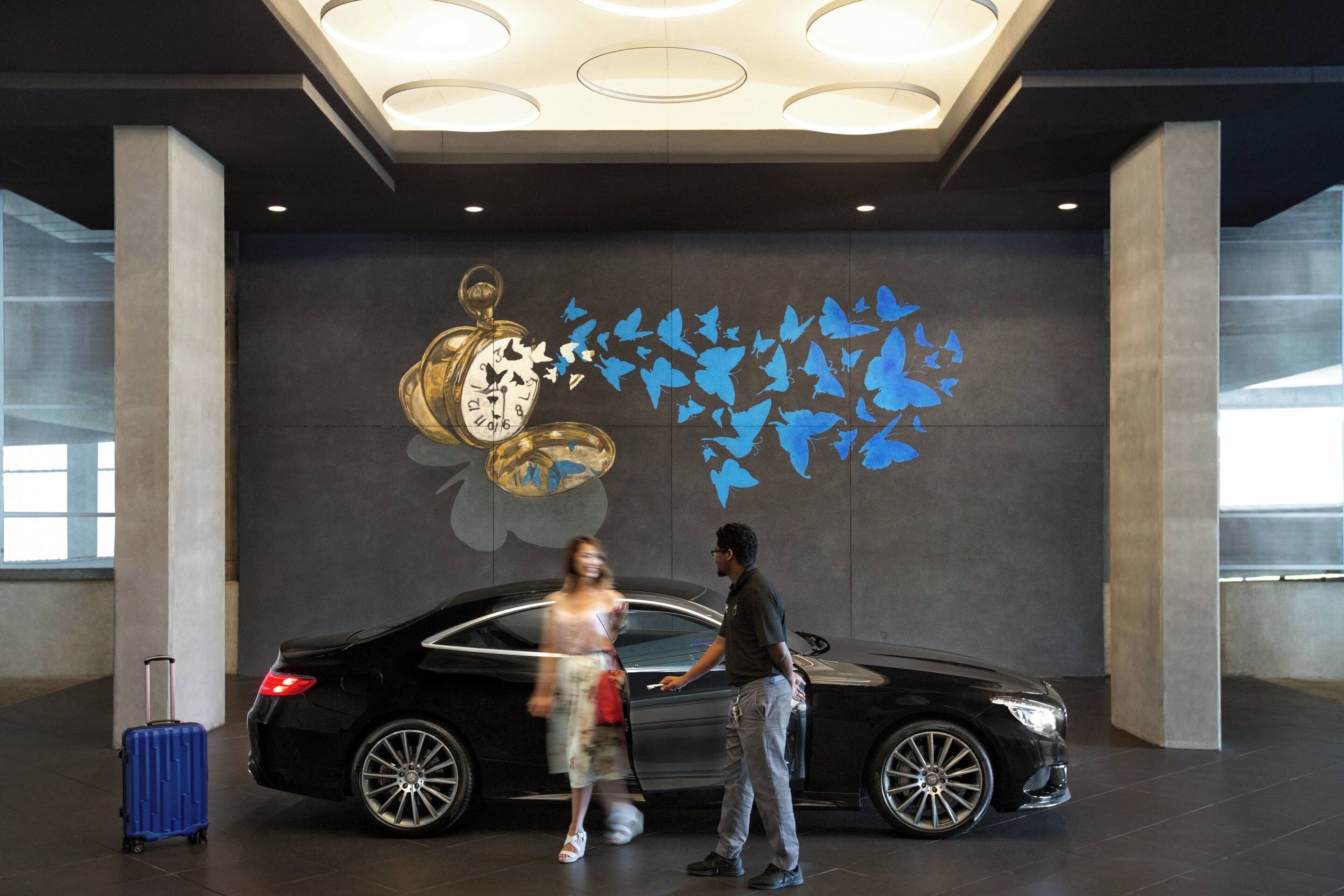 A black car in a spacious lobby with a large wall mural featuring a clock and butterflies.