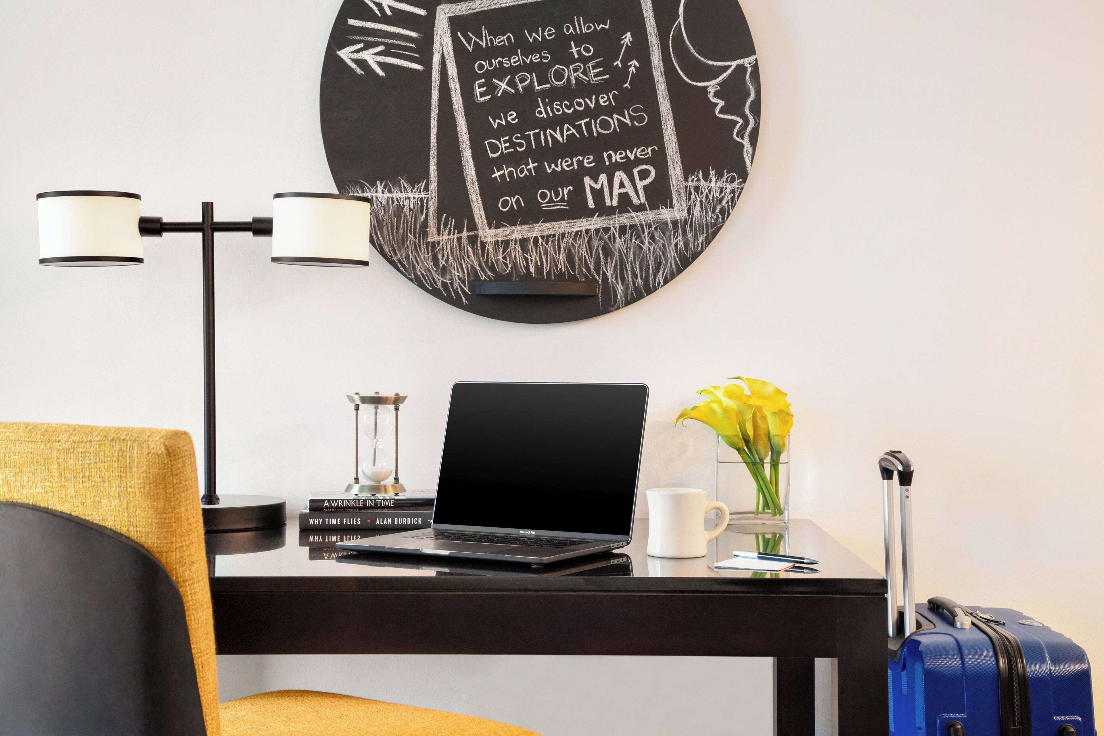 A hotel room with a desk, a laptop, a blue suitcase, a vase with yellow flowers, and a wall art piece with a motivational quote.