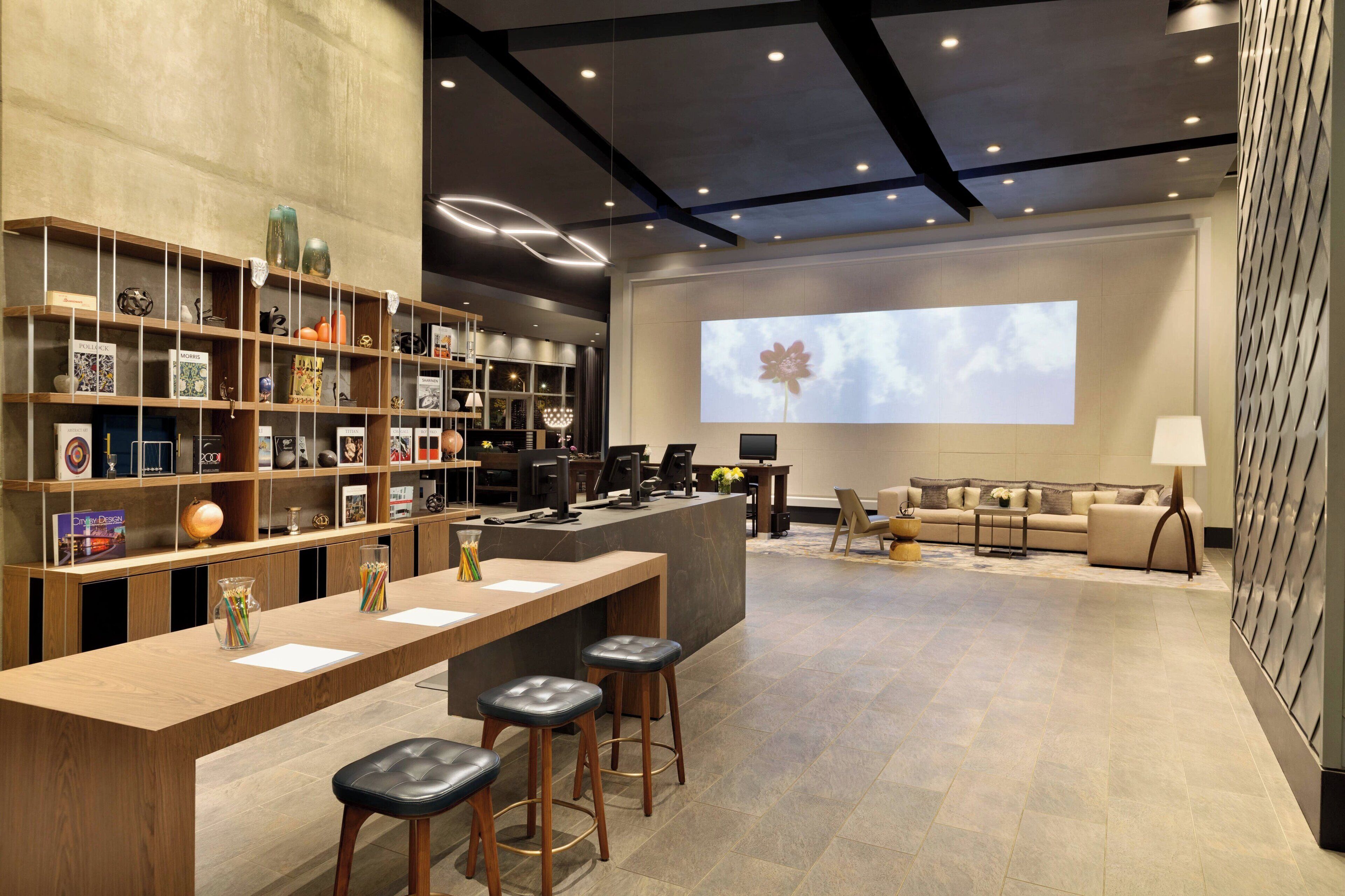 Een moderne hotellobby met een receptiebalie, barstoelen, een bank en een groot scherm waarop een bloem wordt getoond.