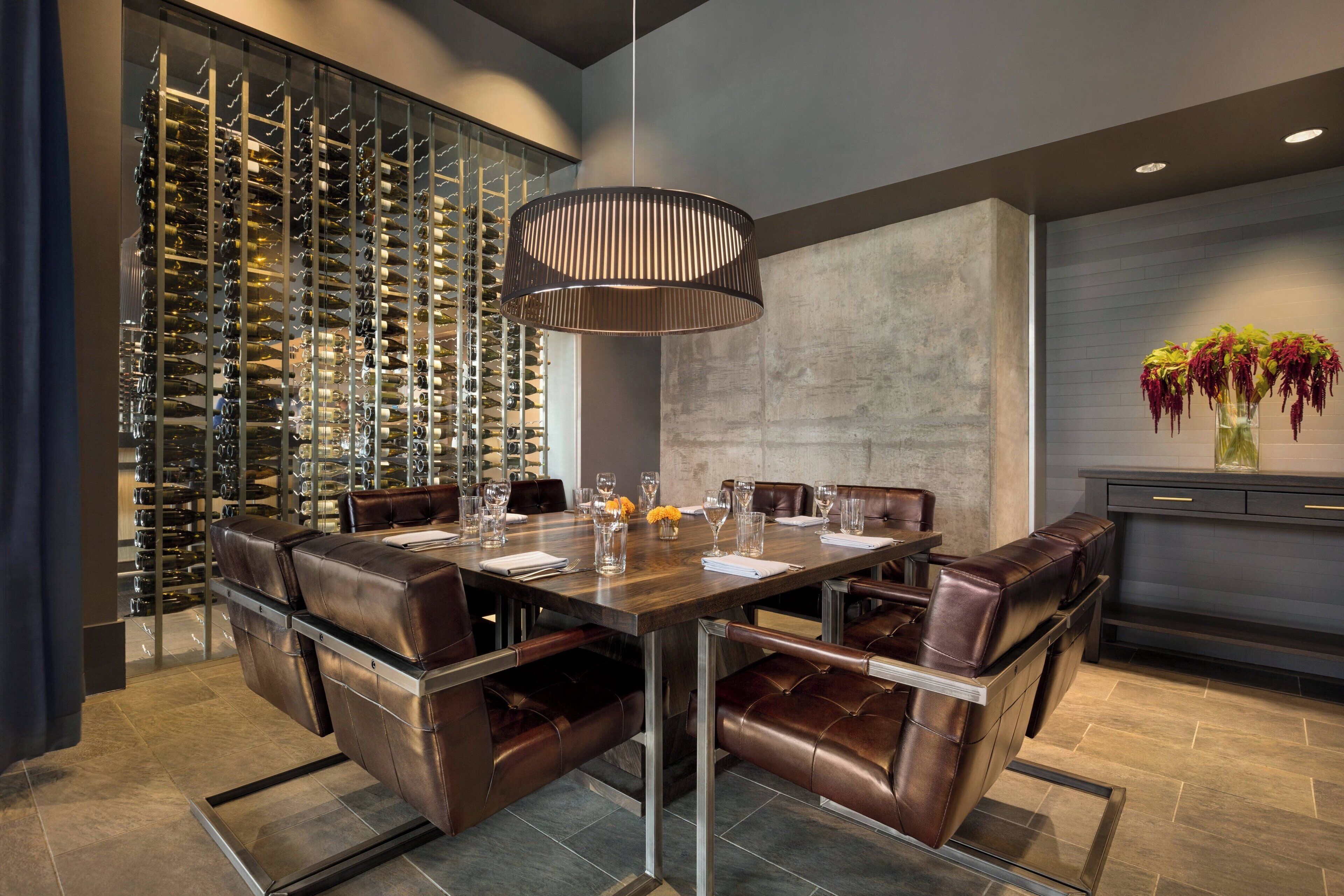 A modern dining area with a wine cellar visible through a glass partition, a central table with chairs, and a decorative vase with flowers on a cabinet.