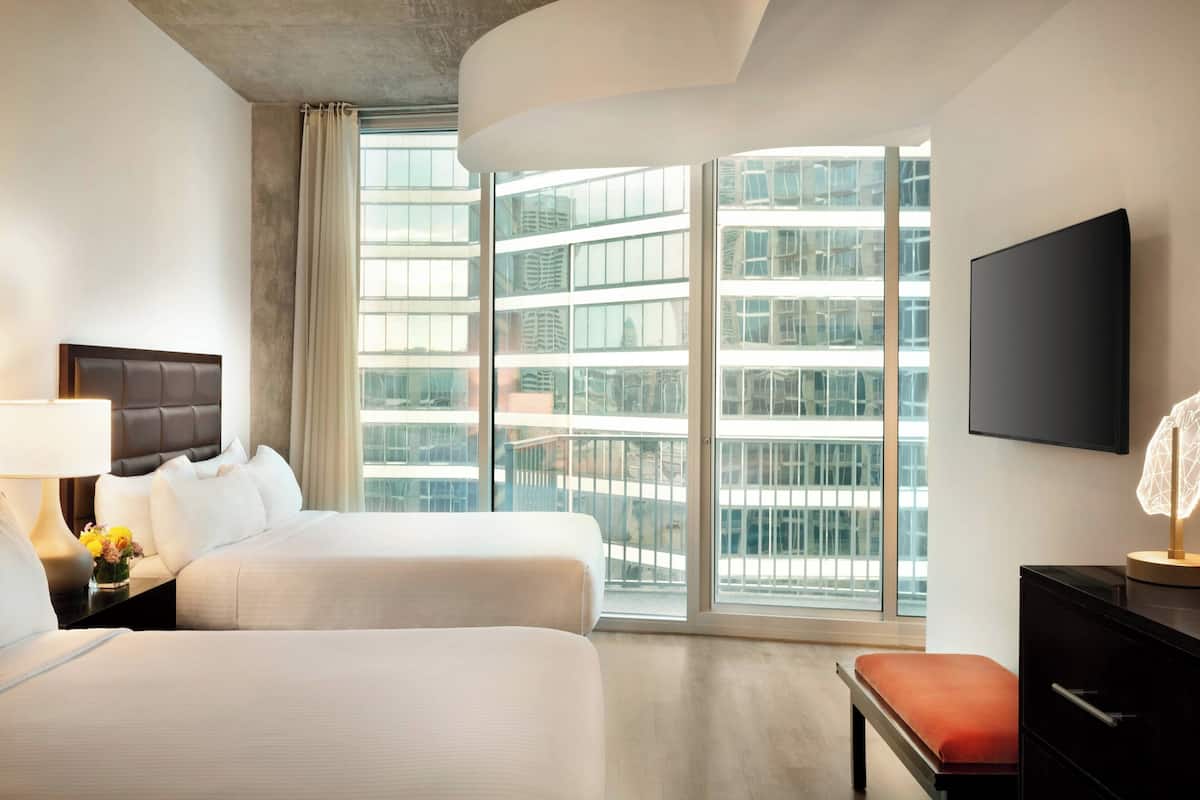 A hotel room with two beds, a TV, a desk, and a large window with a city view.