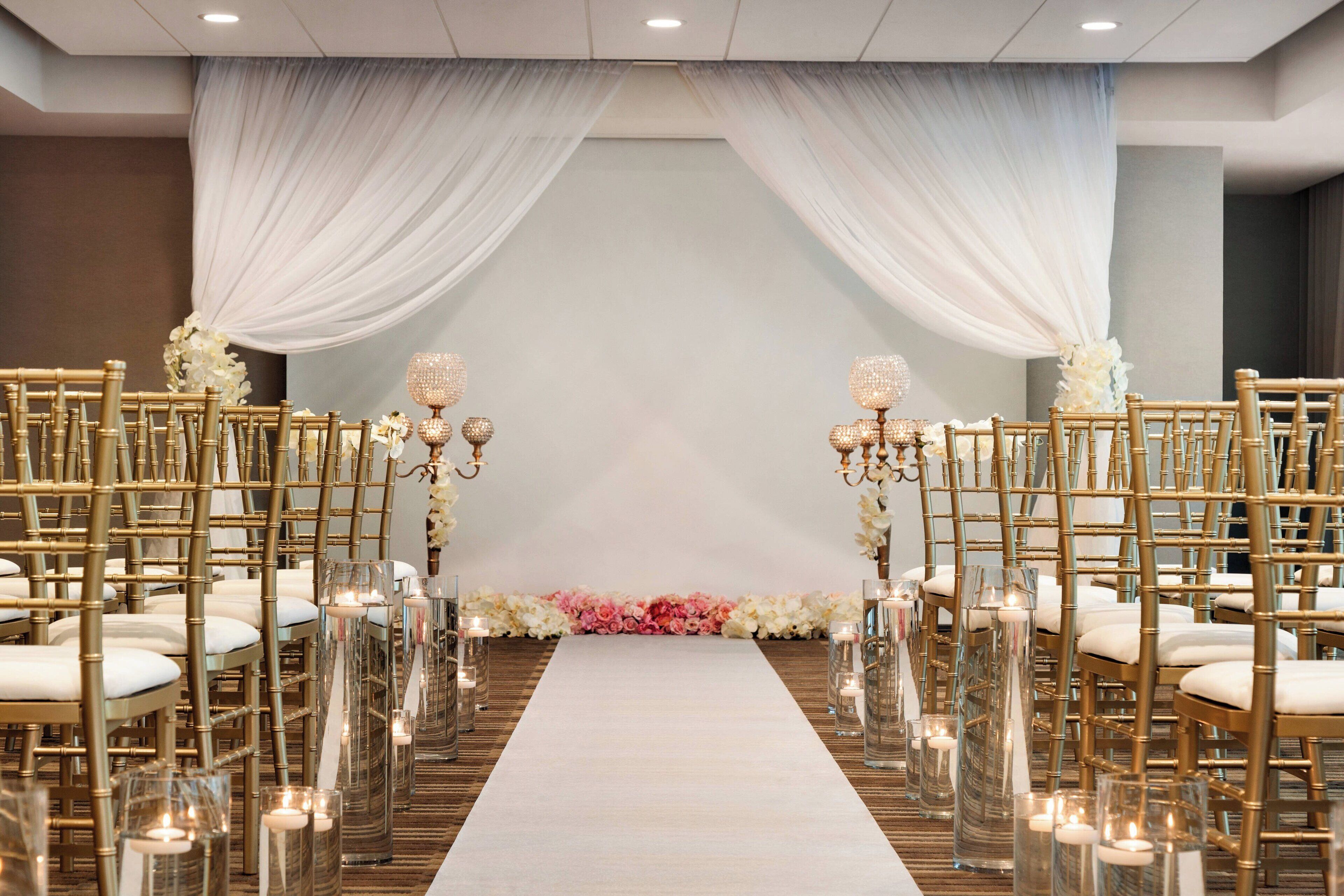 A wedding aisle with white drapes, floral arrangements, and candle-lit tables.