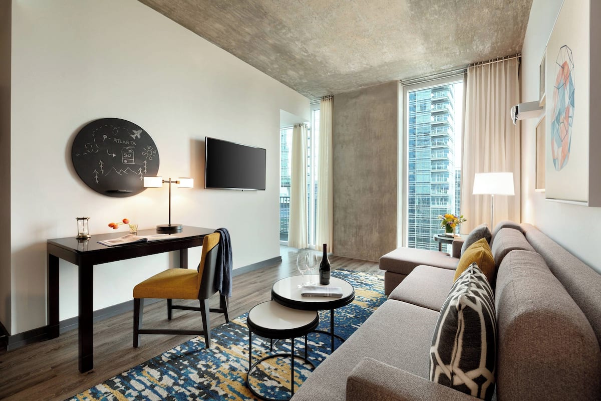 A modern hotel room with a flat-screen TV, a desk with a chair, a sofa, a small round table with a bottle and glasses, and a large window with a city view.