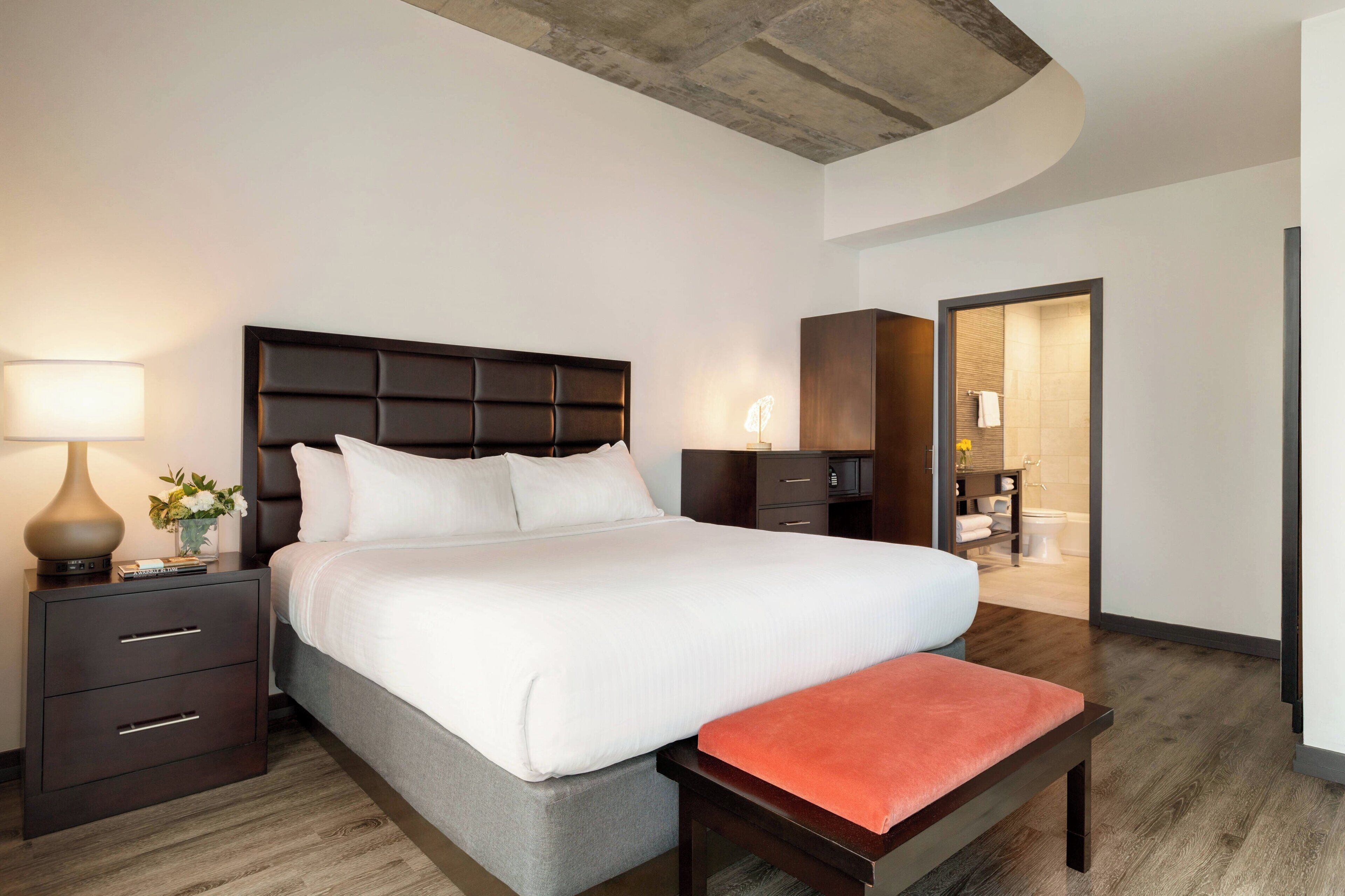 Una habitación de hotel moderna con una cama grande, una mesita de noche con lámpara, un banco y vistas al baño.