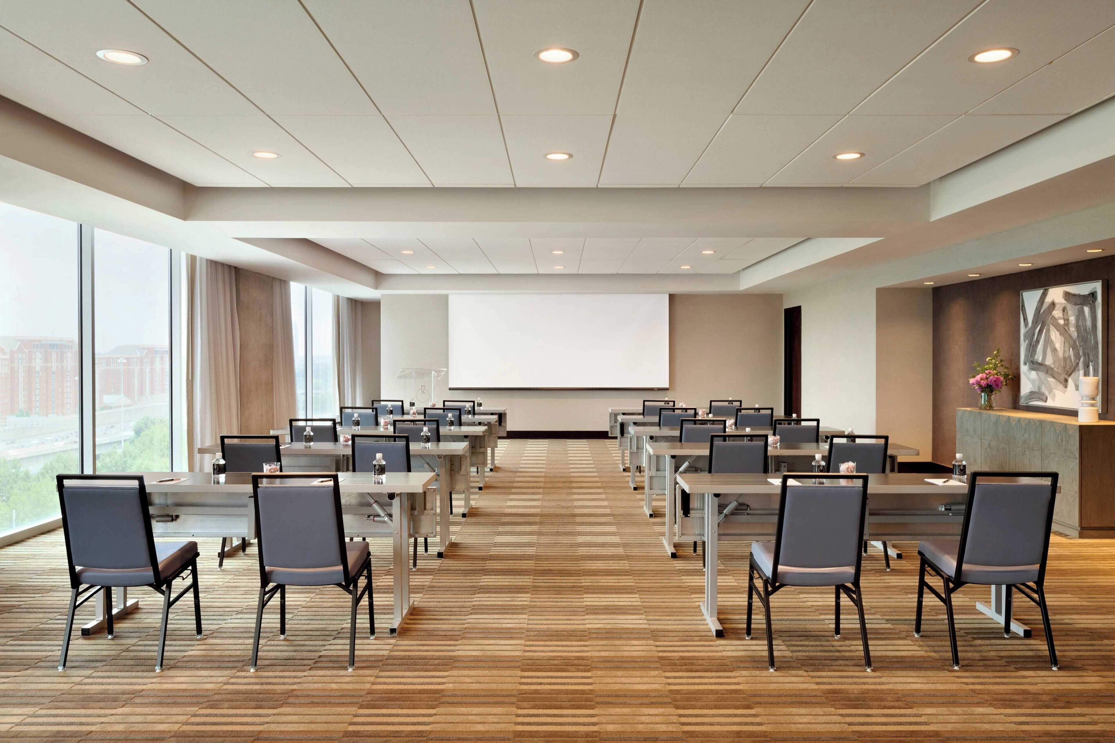 A conference room with multiple tables and chairs, a large screen at the front, and large windows offering a view of buildings outside.