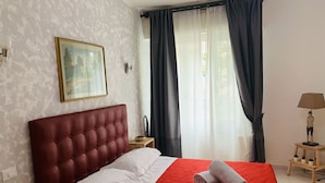 Una cama bien hecha con colcha roja y almohadas blancas, cabecera tapizada en rojo, un cuadro enmarcado en la pared y una ventana con cortinas oscuras.