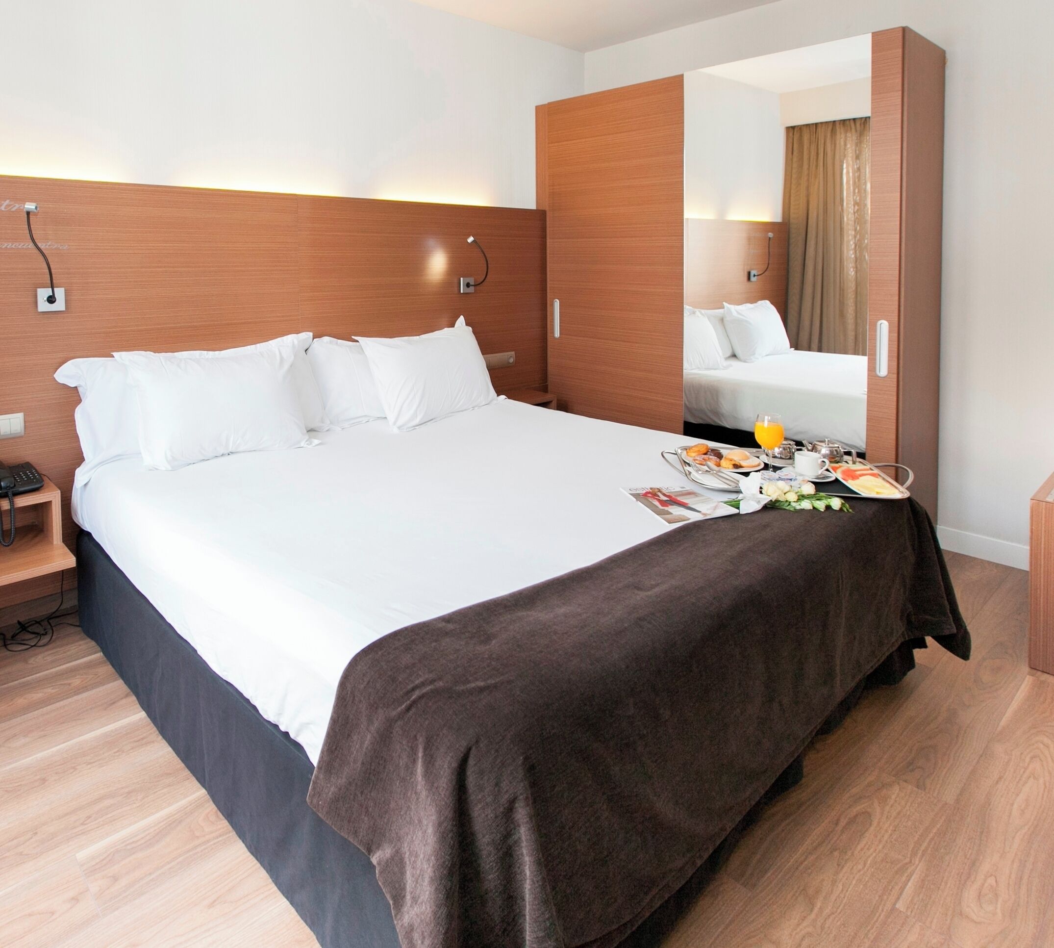 A modern hotel room with a large bed, a bedside table with a phone, a mirror, and a breakfast tray on the bed.
