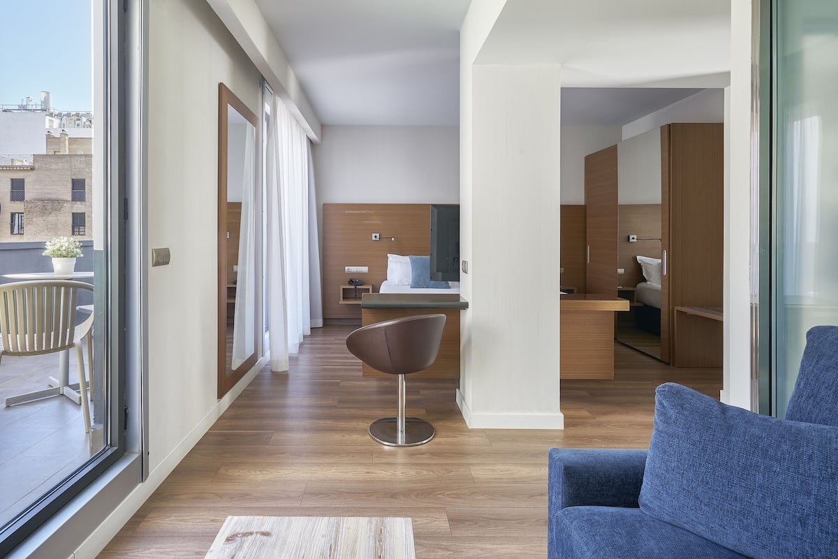 Een moderne hotelkamer met een groot raam, een houten vloer, een blauwe bank, een moderne stoel en een kitchenette op de achtergrond.
