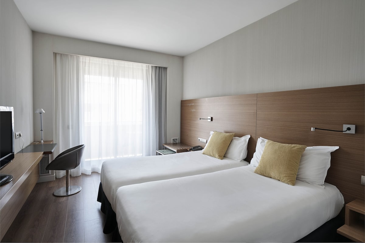 Een hotelkamer met twee bedden, een bureau met stoel, een televisie en een groot raam met gordijnen.