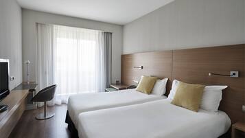 Habitación de hotel con dos camas, un escritorio con silla, televisión y una ventana grande con cortinas.