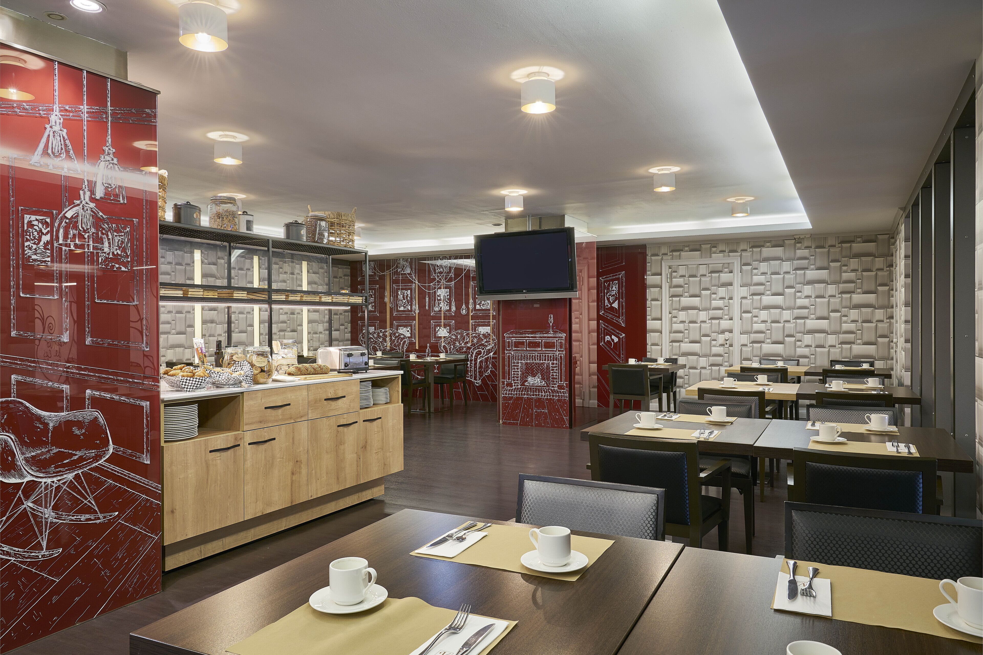 Área de refeições de um hotel moderno, com mesas de madeira, detalhes decorativos em vermelho e uma ilha central com eletrodomésticos de cozinha.