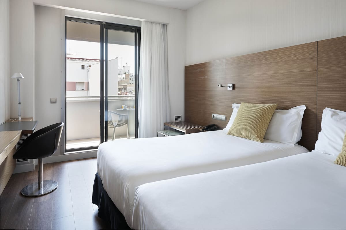 Een hotelkamer met twee bedden, een bureau, een stoel, een klein tafeltje en uitzicht op de stad door een schuifdeur.