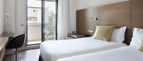 A hotel room with two beds, a desk, a chair, a small table, and a view of the city through a sliding door.