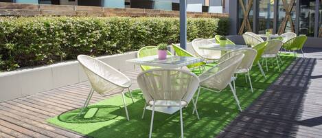 Un patio al aire libre con sillas y mesas de mimbre blanco, rodeado por una terraza de madera y césped artificial verde.