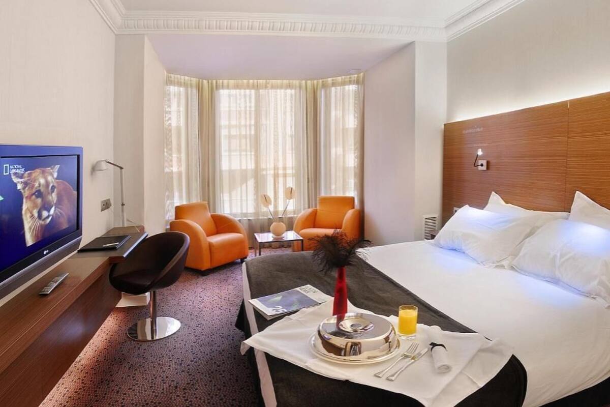 A hotel room with a bed, a TV, a desk with a chair, a table with a fruit bowl, and a window with curtains.