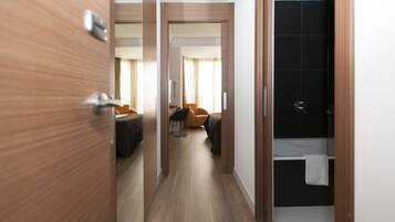 Habitación de hotel con piso de madera, cama con cubrecama oscuro, una silla naranja, escritorio con lámpara y baño con pared de azulejos negros y toalla blanca colgada en la puerta.