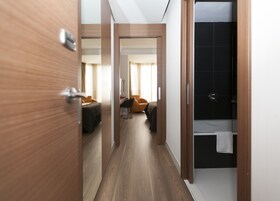 Ein Hotelzimmer mit Holzboden, einem Bett mit dunkelgrüner Bettdecke, einem orangefarbenen Sessel, einem Schreibtisch mit Lampe sowie einem Badezimmer mit schwarzem Fliesenbelag und einem weißen Handtuch, das an der Tür hängt.